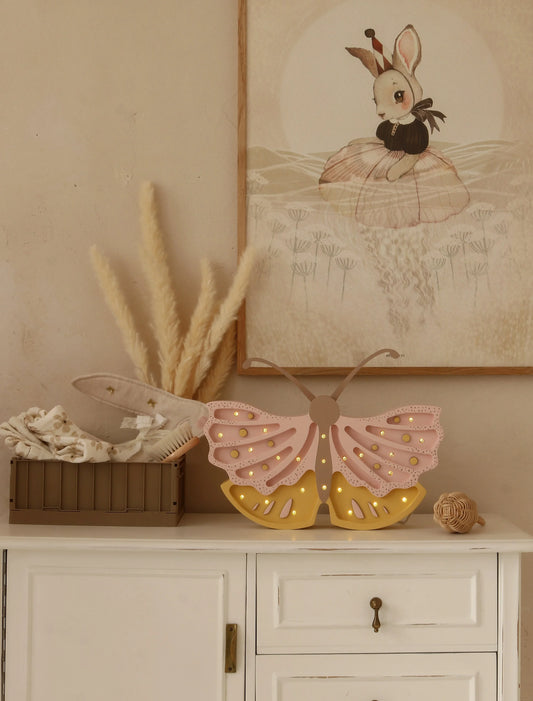 Decorative butterfly lamp on a shelf with a framed picture of a rabbit in a basket in the background.
