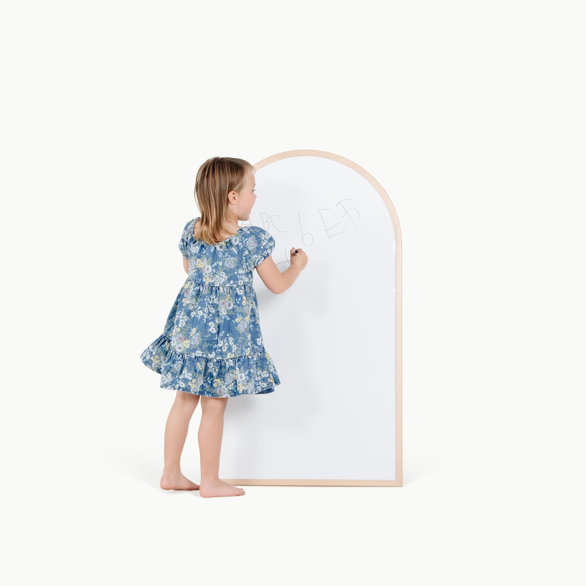 Child in a blue floral dress standing next to a whiteboard with a wooden frame on a white background