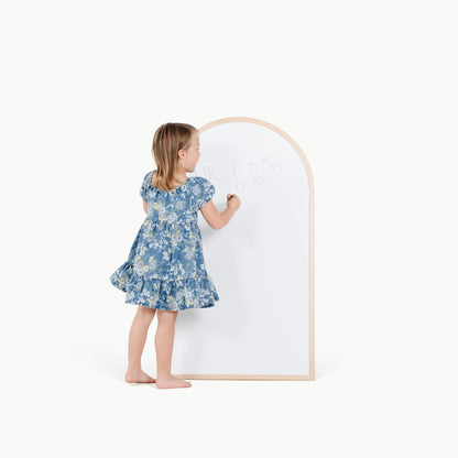 Child in a blue floral dress standing next to a whiteboard with a wooden frame on a white background