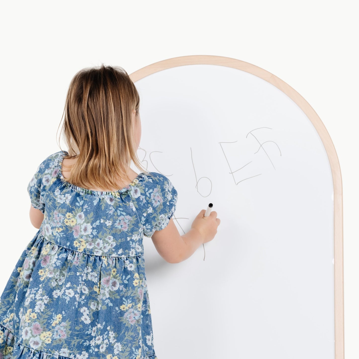 Child in a floral dress writing on a whiteboard with wooden frame.