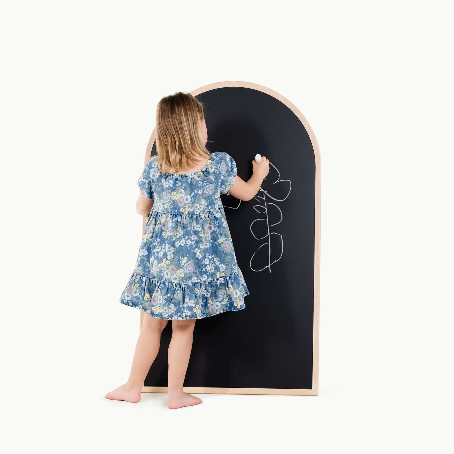 Child in a floral dress writing on a large blackboard with a wooden frame.