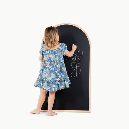 Child in a floral dress writing on a large blackboard with a wooden frame.