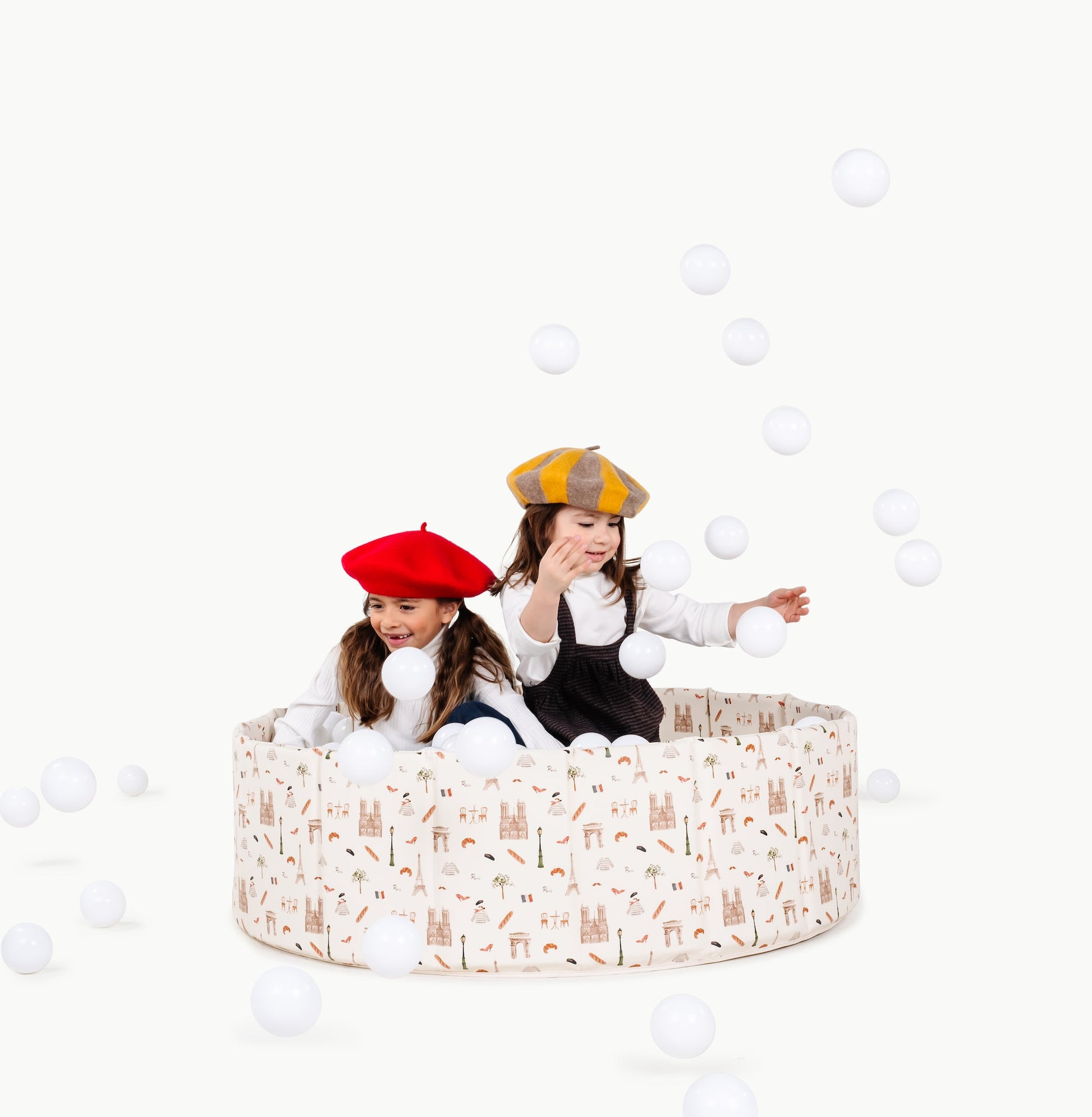 Two children playing in a patterned playpen with white balls scattered around on a white background