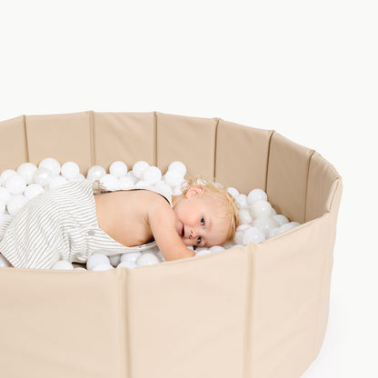 Baby lying in a beige playpen filled with white polystyrene balls on a white background