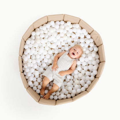 Baby lying on white ball pit balls inside a cardboard box on a white background