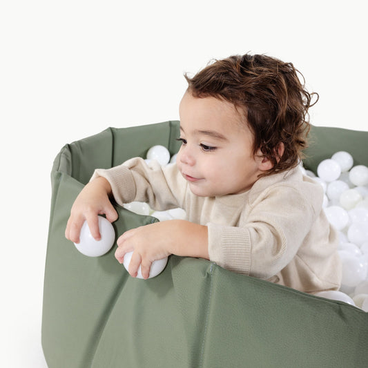 Child playing with white balls inside a green playpen on a white background